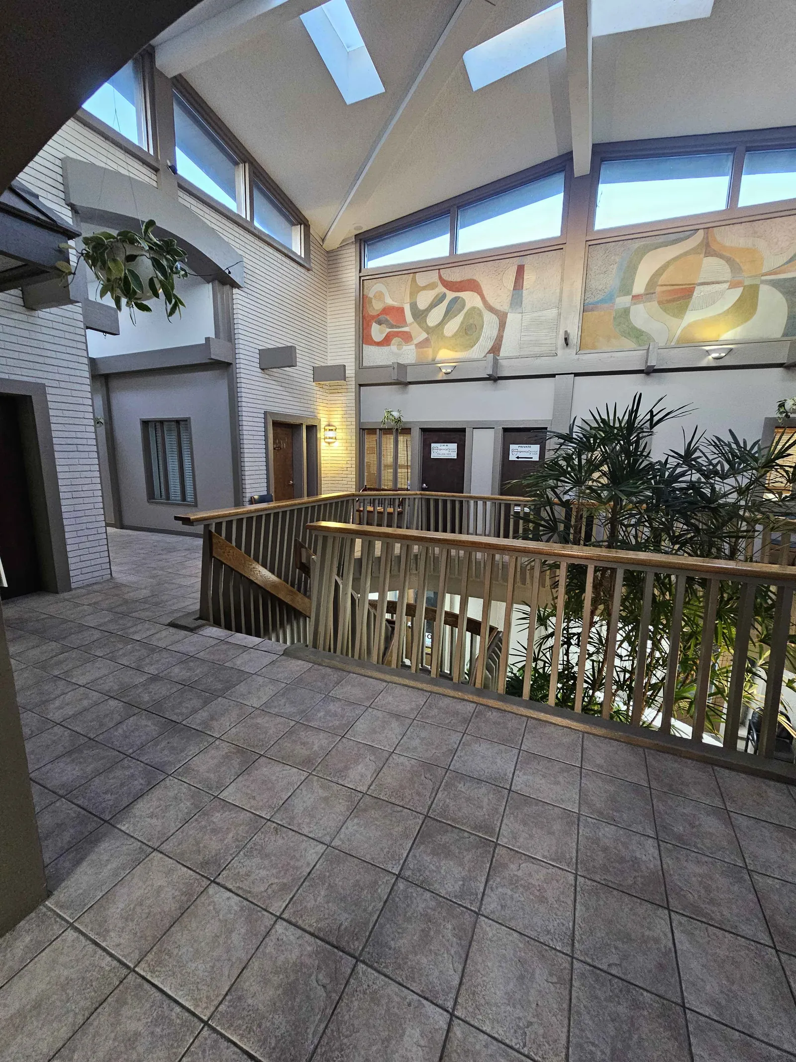 Building atrium lobby at 10000 Watson Rd — skylights, mid-century art mural, staircase, and indoor plants
